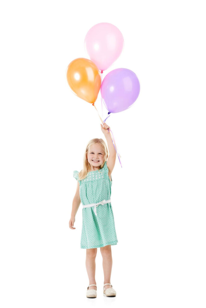 Ein blondes, lachendes Mädchen in einem mintgrünen Sommerkleid hält drei bunte Luftballons (rosa, orange und lila) in der rechten Hand hoch. Sie steht vor weißem Hintergrund und strahlt fröhlich in die Kamera.