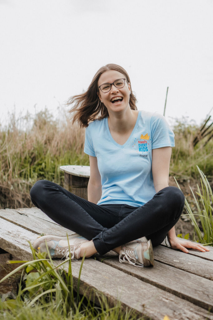 Eine junge Frau mit langen braunen Haaren und Brille sitzt fröhlich lachend im Schneidersitz auf einem hölzernen Steg im Grünen. Sie trägt ein hellblaues T-Shirt mit dem Logo von „Franzi’s Event Kids“ und eine schwarze Hose. Im Hintergrund sind Gräser und Pflanzen zu sehen – die Szene wirkt natürlich und herzlich.