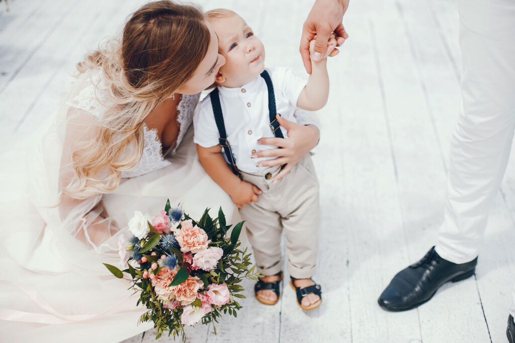 Eine Braut mit langen, lockigen Haaren kniet in ihrem Hochzeitskleid neben einem kleinen Jungen, den sie liebevoll küsst. Der Junge trägt ein weißes Hemd mit Hosenträgern, beige Hosen und Sandalen. Er hält die Hand eines Erwachsenen. Im Vordergrund liegt ein bunter Brautstrauß mit rosa, weißen und blauen Blüten auf dem hellen Holzboden.