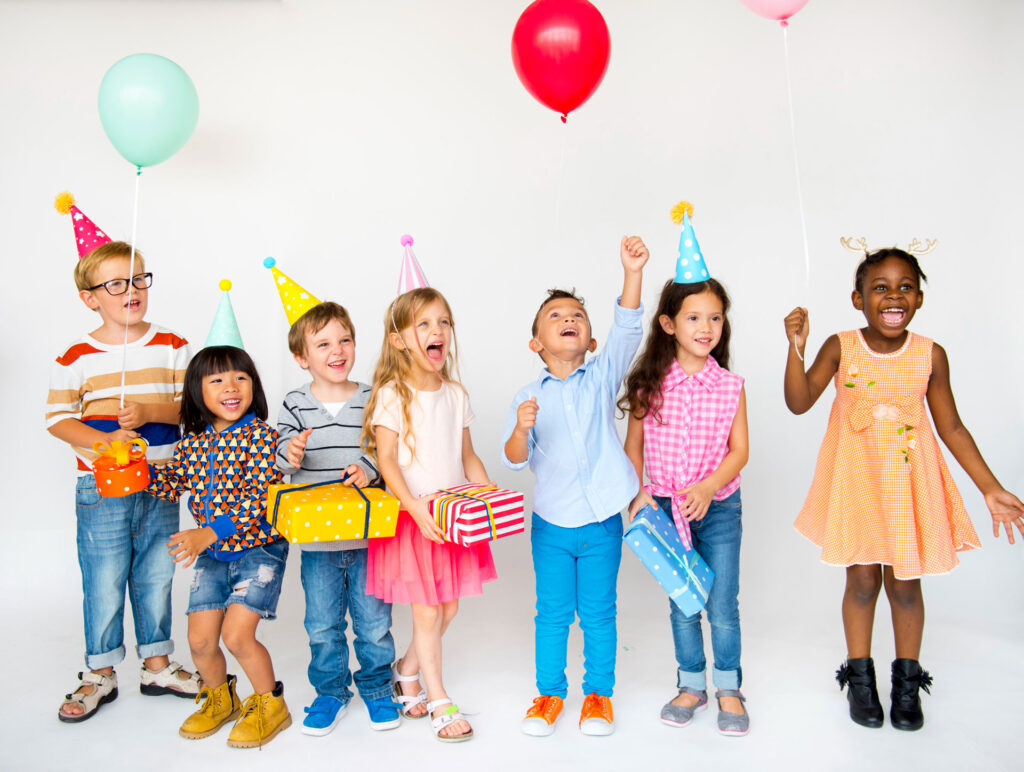 Sieben fröhliche Kinder in Party-Outfits stehen nebeneinander vor weißem Hintergrund. Sie tragen bunte Partyhüte, halten Geschenke und Luftballons in den Händen und lachen oder jubeln ausgelassen. Die Szene strahlt Lebensfreude, Vielfalt und Geburtstagsstimmung aus.