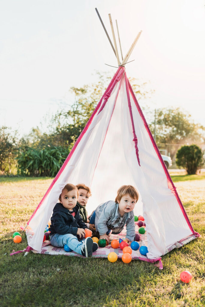 Drei Kleinkinder sitzen in einem weißen Spielzelt mit pinken Rändern, das auf einer Wiese aufgebaut ist. Vor ihnen liegen viele bunte Bälle auf einer Decke. Die Kinder schauen neugierig in die Kamera, die Stimmung ist ruhig und freundlich, das Licht warm und weich.